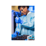 Black female in full PPE is pippetting solution into dark brown test tubes.