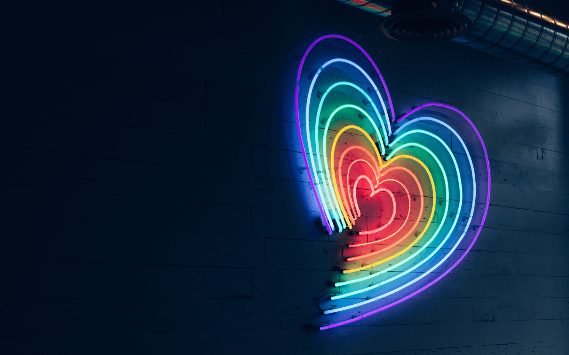 Neon light with expanding heart outline from pink for centre heart to violet on the outermost outline on dark wall. 
