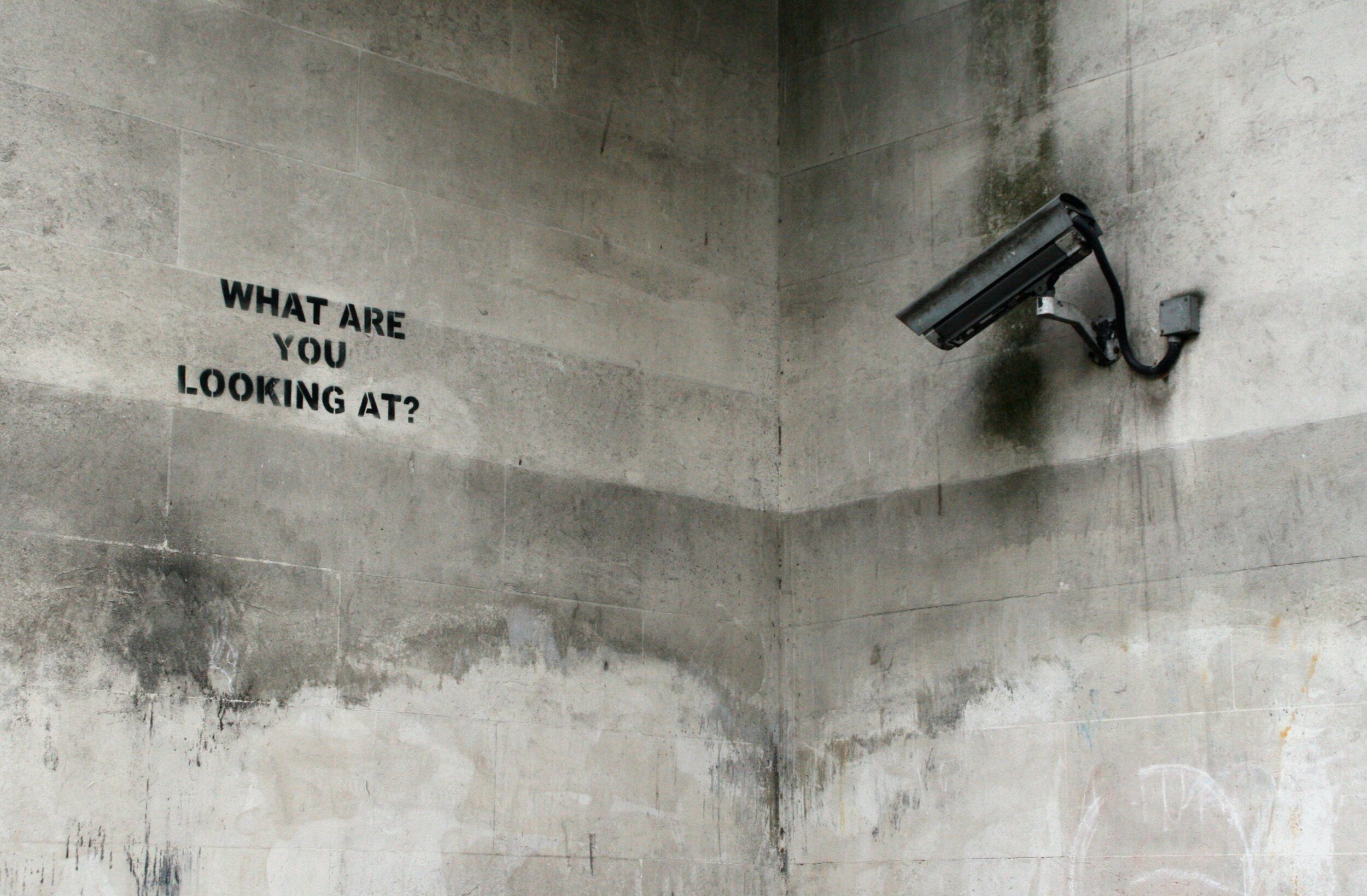 Corner of a concrete wall with CCTV pointing at graffiti saying 'what are you looking at'