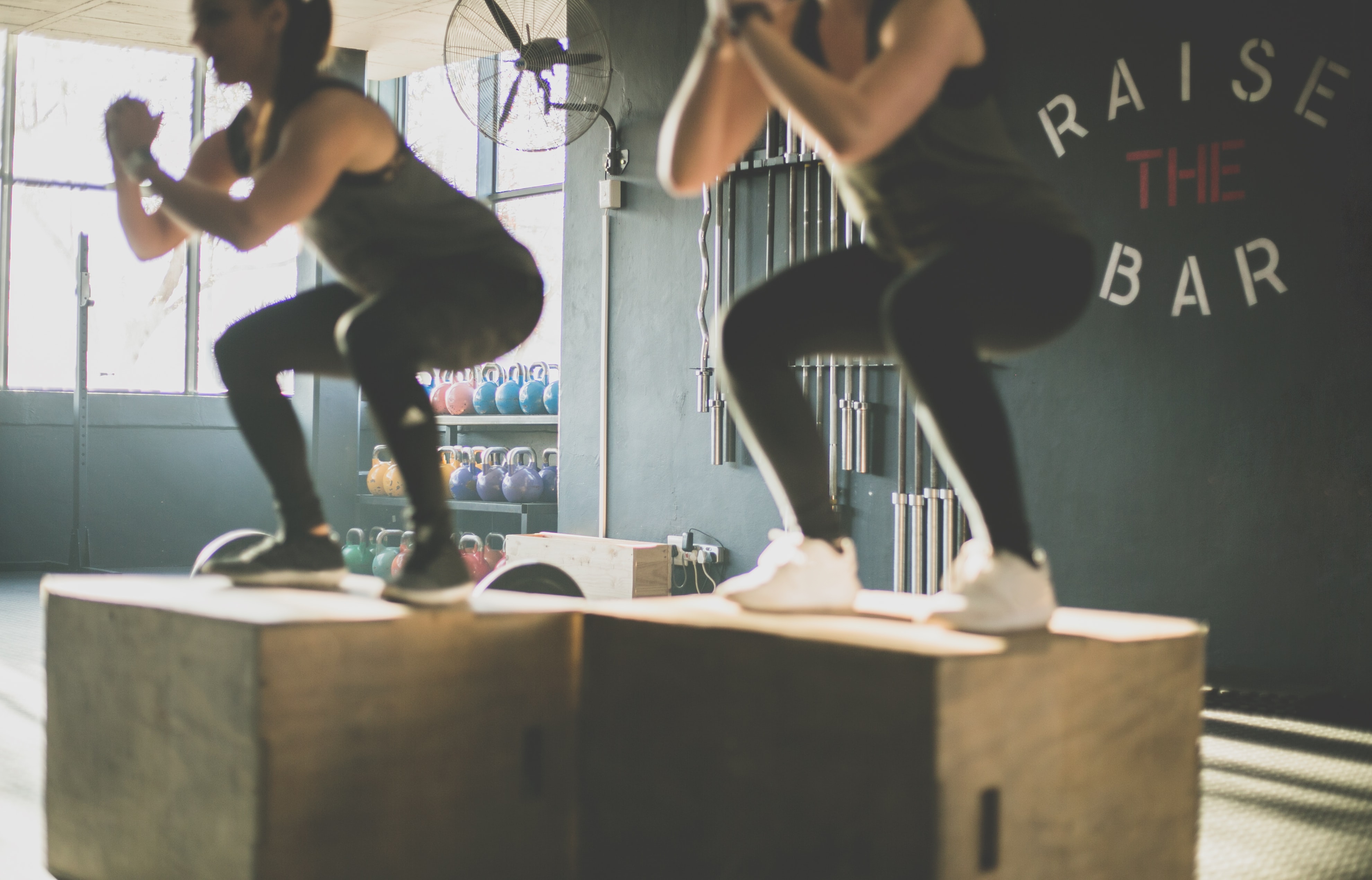 Two women in dark sportswear squatting ontop of boxes in a gym. Raise the bar is written behind them on the wall.