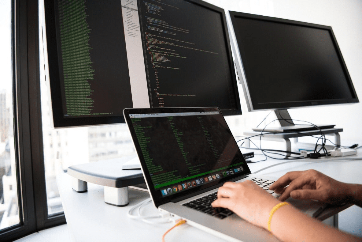 Image of hand typing on a laptop with data filled computer monitors in the background for IT Operations